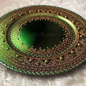May include: An ornate green plate with gold and emerald detailing. The plate has a central dark green section, encircled by elaborate patterns and accents. The text "@mchndibytasha" is visible on the plate.