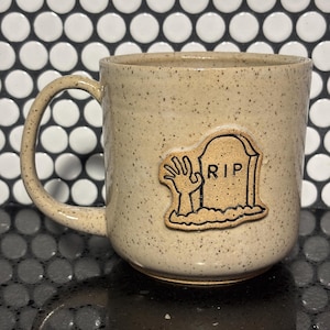 May include: A speckled beige ceramic mug featuring a raised, hand-applied clay applique on the front. The applique depicts a tombstone with the letters RIP engraved on it, accompanied by a hand reaching up from the ground beside the tombstone.