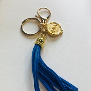 以下が含まれることがあります： 青いタッセルが付いた金のキーホルダー。キーホルダーには、「Ola」という文字が刻まれた丸いチャームが付いています。