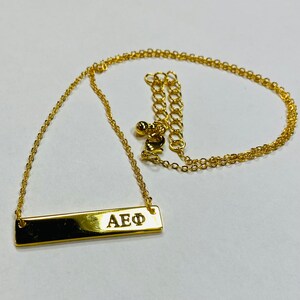 以下が含まれることがあります： 金色のチェーンと小さな金色の留め具が付いた、金色のバーネックレス。バーには「AEΦ」という文字が刻まれています。