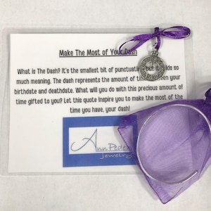 May include: A purple satin bag with a silver bracelet and a silver charm with a clock face and the words "Make the Most of Your Dash". The charm is attached to a purple ribbon. The bag is on a white card with the text "Make The Most of Your Dash" and a quote about the meaning of the dash.