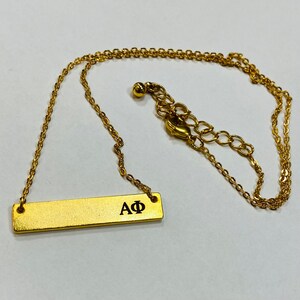 以下が含まれることがあります： 「AΦ」という文字が刻まれた金のバーペンダントが付いた金のネックレスです。