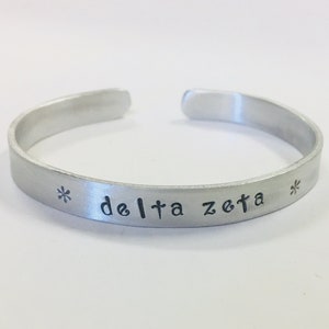 Puede incluir: Una pulsera de puño plateada con las palabras "delta zeta" estampadas en la parte delantera. La pulsera tiene un diseño abierto y está decorada con símbolos de asterisco a cada lado del texto. El metal tiene un acabado cepillado.