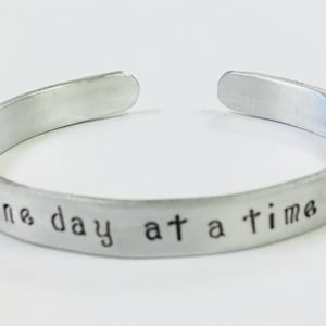 May include: Silver metal bracelet with the inscription "one day at a time".