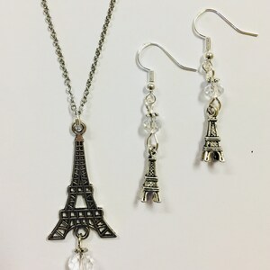 May include: A silver necklace and earrings set featuring a charm of the Eiffel Tower with a crystal pendant. The earrings also feature a crystal and a smaller Eiffel Tower charm.