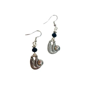 Puede incluir: Un par de pendientes de perezoso plateados. Cada pendiente presenta un pequeño y detallado dije de perezoso colgando de una cuenta negra y un gancho plateado. Los perezosos están representados en una pose relajada.