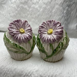 May include: A set of two ceramic salt and pepper shakers with purple flowers and green leaves.