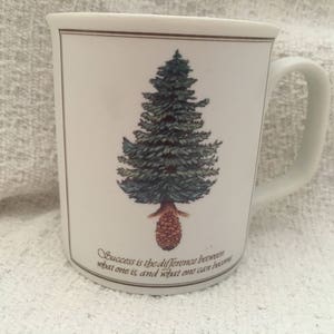 May include: White ceramic mug featuring a detailed illustration of a green and blue pine tree with a brown pinecone base. The mug has a brown border and a quote that reads, "Success is the difference between what one is and what one can become."