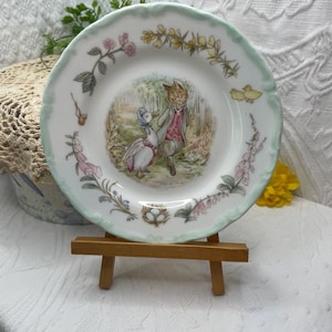 May include: A white decorative plate with a light green scalloped edge. The plate features a central illustration of two anthropomorphic characters in a forest scene, surrounded by floral and bird designs.