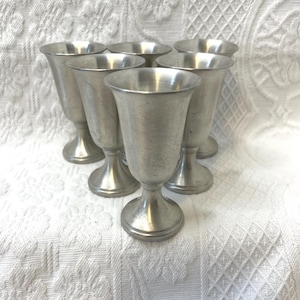 May include: Set of six vintage pewter goblets. Each goblet has a flared rim, a slender body, and a pedestal base. The goblets are arranged in a circular pattern, showcasing their classic design and metallic sheen. The background is a white, textured fabric.