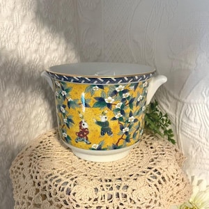 May include: A decorative ceramic planter with a yellow background, featuring a pattern of children, flowers, and leaves. The planter has a white interior, gold trim, and two small handles. The design includes blue and white accents.