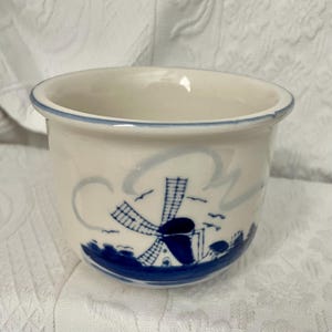 May include: A white ceramic planter with a blue rim and a blue and white windmill design. The planter has a cylindrical shape and is decorated with a Dutch windmill scene. The design includes a windmill, clouds, and birds.