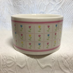 May include: A white ceramic bowl with a pink band at the top and bottom. The bowl is decorated with a repeating pattern of colorful tulips in pink, yellow, and blue.