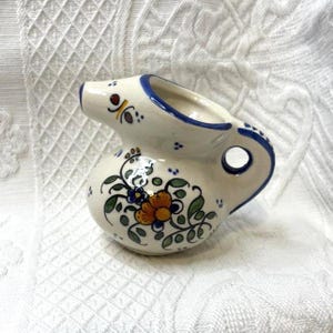 May include: A small, white ceramic pitcher with a blue rim and handle. The pitcher is decorated with a floral design in orange, blue, and green. It has a spout and a round hole in the handle.
