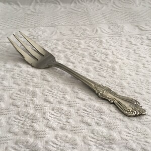 May include: A silver fork with three tines and an ornate handle. The fork is made of metal and rests on a white, textured surface. The handle features a decorative floral design.