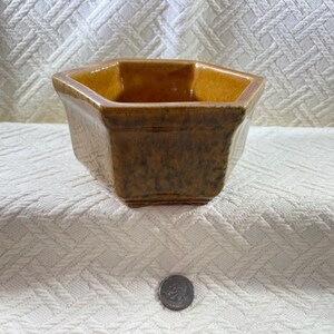 May include: A hexagonal ceramic planter with a speckled brown and tan glaze. The planter has a slightly textured surface and a square base. The interior is a lighter shade of brown. A coin is in the foreground.