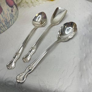 May include: Three silver serving spoons with ornate handles. The spoons have different bowl shapes, including a round, a scoop, and a scalloped design. The spoons are arranged on a white, textured surface.