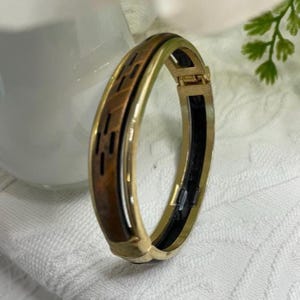 May include: A gold-toned hinged bangle bracelet with a rectangular design. The bracelet features a copper-colored center section with a series of rectangular cutouts. The interior is black. The bracelet is on a white surface.