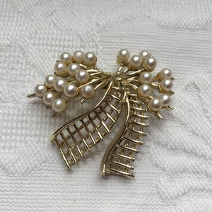 May include: A gold-toned brooch featuring a bow design with a cluster of faux pearls. The bow has a lattice pattern on the ribbon. The brooch is a vintage accessory, suitable for adding a touch of elegance to any outfit.