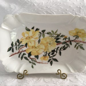 May include: A rectangular white porcelain serving dish with scalloped edges, featuring a central botanical illustration of yellow roses with green leaves and thorny brown stems, displayed on a small gold-toned metal wire stand.