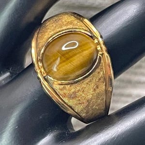 May include: A gold-toned ring featuring an oval tiger's eye stone. The ring has a textured band and a detailed setting around the stone. The tiger's eye stone has a warm, brown color with natural patterns.