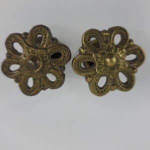May include: Two ornate brass drawer pulls in the shape of a flower with a circular center. The pulls have a detailed design with intricate cutouts.