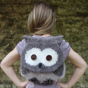 Pode incluir: Uma mochila cinza em forma de coruja com olhos de botão castanhos. A mochila é feita de um material macio e felpudo e está sendo usada por uma pessoa com cabelo castanho comprido.