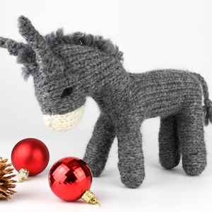 May include: A gray knitted donkey toy with a white nose and a long tail. The donkey is standing on a white surface with three red Christmas ornaments.