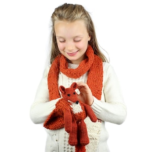 May include: A young girl wearing a white sweater and an orange knitted scarf holds a knitted orange fox toy.
