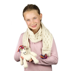 May include: A young person wearing a pink jumper and a white knitted scarf. They are holding a white knitted unicorn toy with colourful yarn hair.