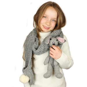 May include: A young girl wearing a white sweater and a gray knitted scarf with a white pom-pom. She is holding a gray knitted bunny toy with pink ears.