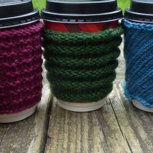 May include: Five coffee cups with different colored knitted cozy covers. The covers are yellow, maroon, green, blue, and brown.