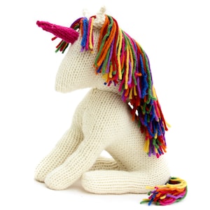 May include: A white knitted unicorn toy with a rainbow mane and tail. The unicorn is sitting on its hind legs with its head turned to the side.