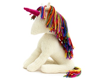 May include: A white knitted unicorn toy with a rainbow mane and tail. The unicorn is sitting on its hind legs with its head turned to the side.