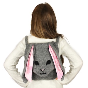May include: A gray felt bunny backpack with pink felt ears. The backpack is worn by a person with long brown hair.