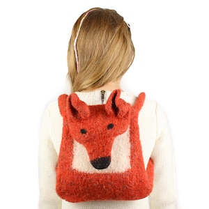 May include: A red fox-shaped backpack made of felt with a white face and black eyes. The backpack is being worn by a person with long blonde hair.
