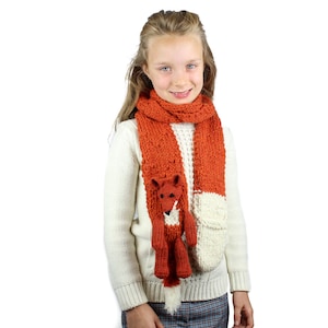 May include: A girl wearing a white sweater and a knitted orange and white scarf with a fox attached. The fox has a white belly and a black nose.