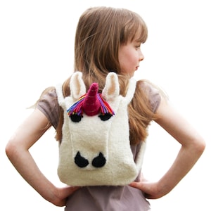 May include: A white knitted unicorn backpack with a pink horn and rainbow mane. The backpack is being worn by a young girl with long brown hair.