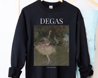 May include: A navy blue sweatshirt featuring a framed print of Edgar Degas's "The Star" ballet painting. The word "DEGAS" is printed in white above the artwork. The sweatshirt is displayed on a wooden hanger with light blue jeans.