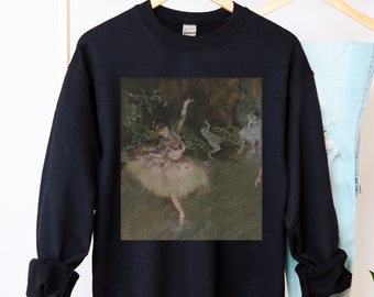 May include: Black sweatshirt with a print of a painting of a ballerina in a white tutu and pink bodice. The ballerina is dancing on a stage with a backdrop of trees and foliage.