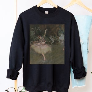 May include: Black sweatshirt with a print of a painting of a ballerina in a white tutu and pink bodice. The ballerina is dancing on a stage with a backdrop of trees and foliage.