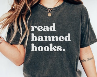 May include: A woman wearing a dark grey t-shirt with the text "read banned books." in white lettering. She is wearing blue jeans and white sneakers.