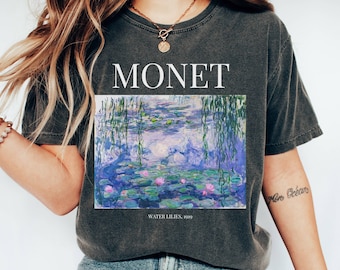May include: A charcoal grey t-shirt with a graphic print of Claude Monet's "Water Lilies" painting. The text "MONET" is printed above the painting and "WATER LILIES, 1919" is printed below the painting.