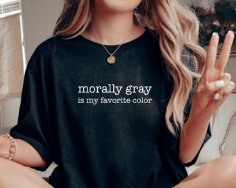 May include: A person wearing a black t-shirt with the text "morally gray is my favorite color" in white. The person is sitting on a bed with a blanket and a tray with a bowl of cereal and a mug.