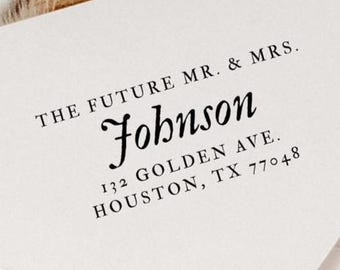 May include: White envelope with the text "THE FUTURE MR. & MRS. Johnson" and address details: 132 GOLDEN AVE. HOUSTON, TX 77048. The envelope is on a wooden surface.
