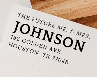 May include: A white card with the text "THE FUTURE MR. & MRS. JOHNSON" and address details in black font. The card is on a wooden surface, with a white envelope in the background. The address is 132 Golden Ave., Houston, TX 77048.