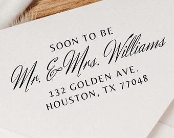 May include: A white envelope with the text "SOON TO BE Mr. & Mrs. Williams" printed in black script. The address "132 GOLDEN AVE. HOUSTON, TX 77048" is also printed on the envelope. The envelope is on a wooden surface.