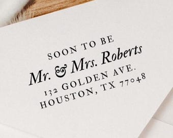 May include: White envelope with the text "SOON TO BE Mr. & Mrs. Roberts" in black. The address is 132 GOLDEN AVE., HOUSTON, TX 77048. The envelope is on a wooden surface.