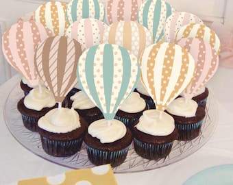 May include: A collection of chocolate cupcakes topped with white frosting and decorative hot air balloon toppers. The toppers feature patterns in pastel colors like pink, blue, and tan, creating a festive and whimsical display.