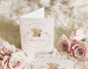 May include: Two white thank you cards with a gold chalice and wheat design. The text on the card reads "May the Lord bless you" and "Thank You". The cards are on a white fluffy surface with pink roses.